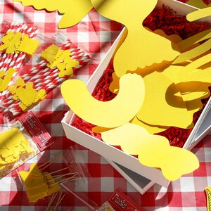 May include: A picnic-themed arrangement on a red and white checkered tablecloth. The scene includes yellow paper cutouts, red and white striped straws, and small packages with text. A white box contains yellow paper shapes and red filler.