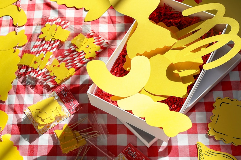 May include: A picnic-themed arrangement on a red and white checkered tablecloth. The scene includes yellow paper cutouts, red and white striped straws, and small packages with text. A white box contains yellow paper shapes and red filler.