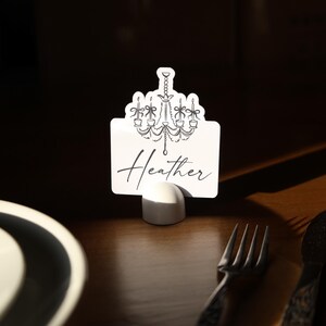 May include: A white place card with a chandelier design and the name "Heather" in script. The card is held in a white holder, set on a wooden table with a fork and knife.