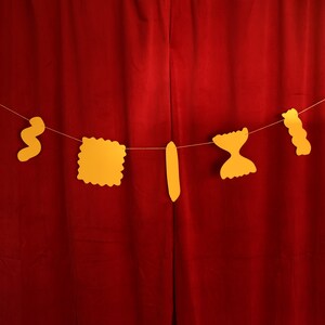 May include: A string of yellow pasta-shaped cutouts hangs against a red curtain. The pasta shapes include elbow macaroni, rotini, a square ravioli, a long noodle, farfalle, and a wavy noodle. The shapes are strung on a thin white string.