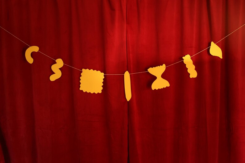 May include: A string of yellow pasta-shaped cutouts hangs against a red curtain. The pasta shapes include elbow macaroni, rotini, a square ravioli, a long noodle, farfalle, and a wavy noodle. The shapes are strung on a thin white string.