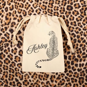 May include: A cream-colored drawstring bag with a black illustration of a leopard and the name "Ashley" in cursive. The bag is set against a leopard print background.