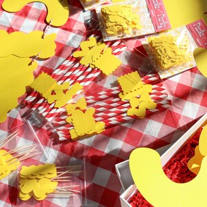 May include: A picnic-themed arrangement on a red and white checkered tablecloth. The scene includes yellow paper cutouts, red and white striped straws, and small packages of yellow confetti. Some items are in a white box with red filler.