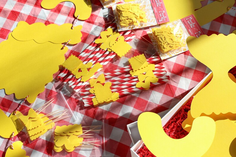 May include: A picnic-themed arrangement on a red and white checkered tablecloth. The scene includes yellow paper cutouts, red and white striped straws, and small packages of yellow confetti. Some items are in a white box with red filler.