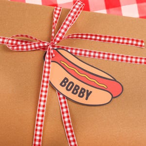 May include: A brown gift box tied with a red and white checkered ribbon. A hot dog-shaped tag with the name "BOBBY" is attached to the ribbon, suggesting a themed gift.