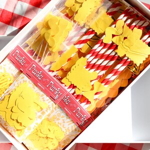 May include: A white box filled with pasta-themed party supplies. Inside are yellow pasta-shaped confetti, red and white striped straws, and packages with the text "Pasta Party". The box sits on a red and white checkered tablecloth.