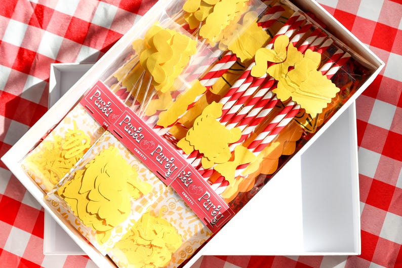 May include: A white box filled with pasta-themed party supplies. Inside are yellow pasta-shaped confetti, red and white striped straws, and packages with the text "Pasta Party". The box sits on a red and white checkered tablecloth.