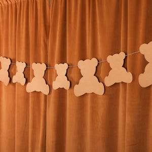 May include: A string of light brown teddy bear cutouts strung on a white string, hanging against a backdrop of a brown curtain. The bears are in various sizes and shapes, creating a festive decoration.