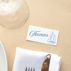 May include: A place setting with a white name card that reads "Thomas" in blue script, with a sailboat illustration. The card is on a tan surface, next to a folded white napkin, silverware, and a wine glass.