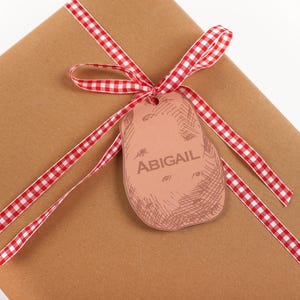 May include: A wrapped gift in brown paper with a red and white checkered ribbon. A potato-shaped gift tag with the name "ABIGAIL" is attached to the ribbon. The gift is on a white background.