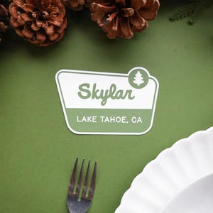 May include: A white and green sticker with the name "Skylar" and "Lake Tahoe, CA" printed on it. The sticker has a tree icon and is on a green surface, with pine cones and a fork in the background.
