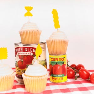 May include: Cupcakes with white frosting and yellow toppers sit atop cans of tomato products. Fresh red cherry tomatoes are scattered on a red and white checkered tablecloth. The cans include Muir Glen Organic crushed tomatoes and Cento Tomato Paste.