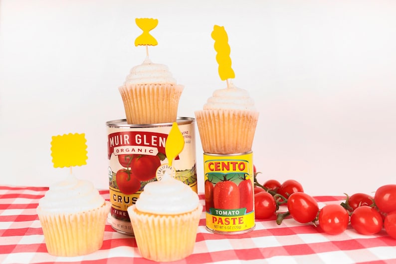May include: Cupcakes with white frosting and yellow toppers sit atop cans of tomato products. Fresh red cherry tomatoes are scattered on a red and white checkered tablecloth. The cans include Muir Glen Organic crushed tomatoes and Cento Tomato Paste.