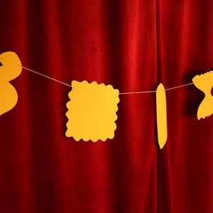 May include: A string of yellow pasta-shaped cutouts hangs against a deep red curtain. The pasta shapes include a wavy noodle, a square with a scalloped edge, a long, flat noodle, a bow tie, and a twisted noodle. The shapes are suspended by a thin, light-colored string.