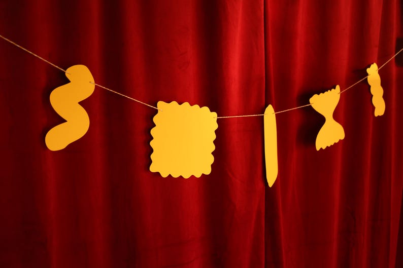 May include: A string of yellow pasta-shaped cutouts hangs against a deep red curtain. The pasta shapes include a wavy noodle, a square with a scalloped edge, a long, flat noodle, a bow tie, and a twisted noodle. The shapes are suspended by a thin, light-colored string.