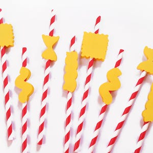 May include: A collection of red and white striped paper straws, each adorned with a yellow pasta-shaped decoration. The pasta shapes include bow ties, wavy ribbons, and squares. The straws are arranged against a white background.