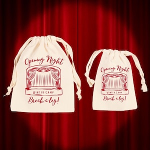 May include: Two off-white drawstring bags with red text and a theater stage graphic. The text reads "Opening Night," "Winter Camp," and "Break a leg!" The bags are on a red curtain background.