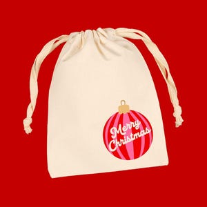 May include: A cream-colored drawstring pouch featuring a graphic of a red and pink striped Christmas ornament. The ornament has a gold cap and the white text Merry Christmas printed across the center in a cursive script font.