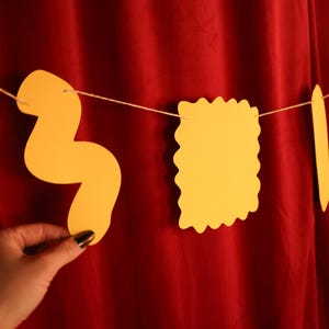 May include: A string of yellow pasta-shaped cutouts hangs against a red backdrop. The shapes include a wavy noodle, a square with a scalloped edge, a long, thin noodle, and a bow tie shape. The pasta shapes are strung on a thin, white string.