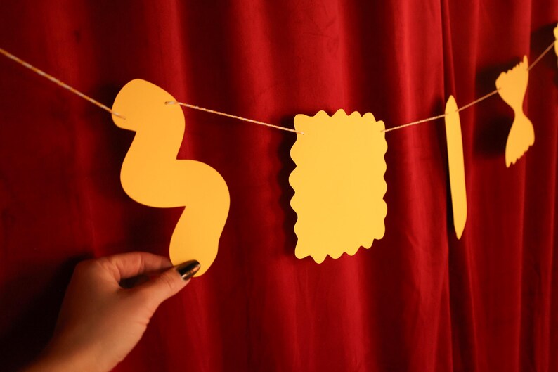May include: A string of yellow pasta-shaped cutouts hangs against a red backdrop. The shapes include a wavy noodle, a square with a scalloped edge, a long, thin noodle, and a bow tie shape. The pasta shapes are strung on a thin, white string.