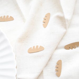May include: A white plate with a scalloped edge sits next to a beige linen cloth. Scattered across the cloth are several small, tan, bread-shaped cutouts. The cutouts have a textured appearance, with lines cut into the top to resemble bread.