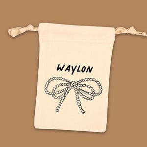 Puede incluir: Una bolsa de cord&oacute;n de color crema con la palabra "WAYLON" en escritura negra sobre una ilustraci&oacute;n de lazo de cuerda negra. Fondo marr&oacute;n.