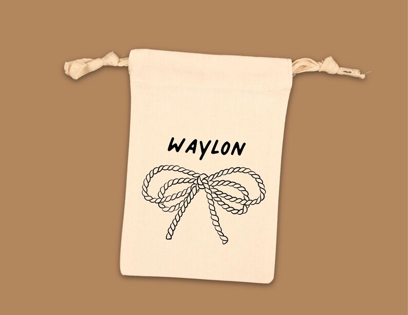 Puede incluir: Una bolsa de cord&oacute;n de color crema con la palabra "WAYLON" en escritura negra sobre una ilustraci&oacute;n de lazo de cuerda negra. Fondo marr&oacute;n.