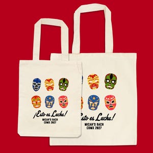 May include: Two off-white tote bags with a red background. Each bag features a colorful design of six luchador masks. The text "¡Esto es Lucha!" and "MICAH'S BACH CDMX 2027" are printed below the masks.