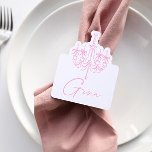 May include: A place setting with a white plate, a pink napkin, and a silver fork. A white place card with a pink chandelier design and the name "Gina" is tucked into the napkin. The setting is on a white surface.