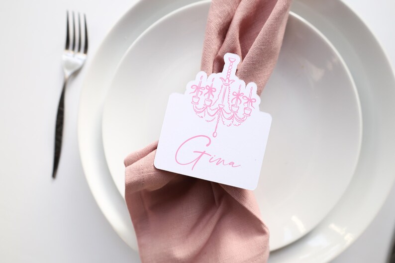 May include: A place setting with a white plate, a pink napkin, and a silver fork. A white place card with a pink chandelier design and the name "Gina" is tucked into the napkin. The setting is on a white surface.