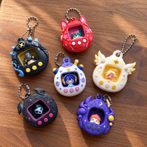 May include: Six digital pet keychain charms in various colors and designs, including black, red, white, purple, and blue, each featuring a small character inside a screen-like window and a metal ball chain attached to the top for hanging.