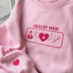 Może przedstawiać: Jasnoróżowa bluza z okrągłym dekoltem i ciemnoróżowym haftem na piersi z napisem HEALER MAIN nad prostokątną ramką zawierającą butelkę z miksturą, skrzydlate serce ze znakiem plus oraz różdżkę. Na rękawie znajduje się małe różowe serce.