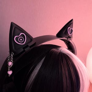 May include: A black headband featuring two pointed cat ears. Each ear is decorated with a light purple heart-shaped emblem. A silver-toned metal chain with two small light purple heart charms hangs from the left ear attachment point.