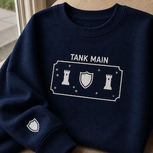 Könnte beinhalten: Ein marineblaues Sweatshirt mit Rundhalsausschnitt und weißer Stickerei. Auf der Brust befindet sich der Schriftzug TANK MAIN über einem rechteckigen Rahmen mit zwei Burgtürmen, einem zentralen Schild und kleinen Sternen. Ein kleiner Schild ist auf den Ärmel gestickt.