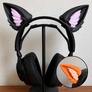 May include: Black over-ear headphones featuring detachable black cat ears with light pink inner accents. A circular inset shows an orange version of the same cat ear attachment. The headphones are mounted on a black stand for display purposes.