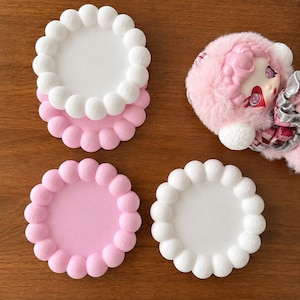 May include: Three circular coasters with scalloped edges featuring a series of rounded spheres along the perimeter. Two coasters are solid white and one is solid light pink. A small doll with pink hair and a fuzzy hood is positioned nearby.