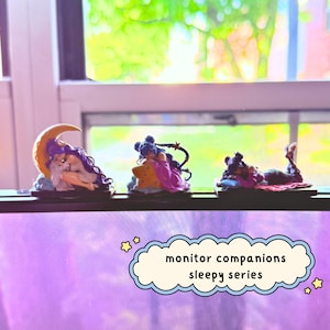May include: Three small figurines from the monitor companions sleepy series. The first features a figure with purple hair resting on a yellow crescent moon. The second shows a figure with dark hair on a star. The third shows a figure on a pillow.