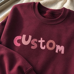 Könnte beinhalten: Ein tiefburgunderfarbenes Sweatshirt mit Rundhalsausschnitt und dem Wort custom, das in verschiedenen Rosa- und Rottönen auf die Brust appliziert ist. Jeder Buchstabe zeigt ein anderes Muster, darunter Vichy-Karo, Punkte und kleine Blumen.