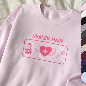 May include: A light pink crewneck sweatshirt featuring the text HEALER MAIN in pink letters. The graphic includes a potion bottle, a winged heart with a plus sign, and a star wand inside a rectangular border, with a vertical color palette strip.