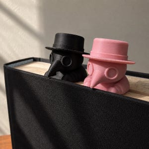 May include: Two plague doctor figurines, one matte black and one light pink, each wearing a top hat and a long beak mask. These small, three-dimensional figures are positioned as book accessories, resting on the top edge of a closed book.