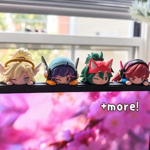 May include: Four small chibi-style figurines resting on a black ledge. The figures feature distinct hair colors including blonde, purple, green, and pink, with various headgear like wings, headphones, and animal ears. The text +more! is visible.
