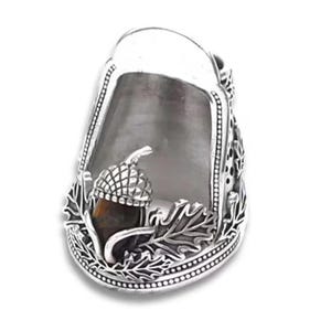 May include: A silver thimble featuring a detailed relief of an acorn with a brown stone inlay, surrounded by sculpted oak leaves. The metal surface has a polished finish with intricate beaded borders along the base and the top opening edge.