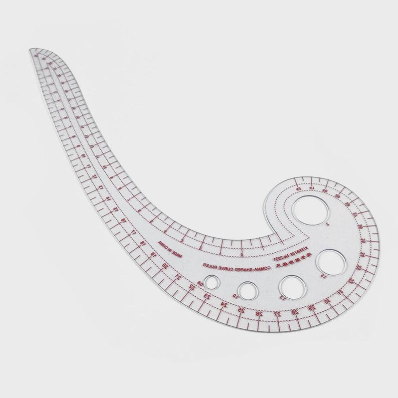 May include: A clear, curved ruler with red markings and measurements in inches. The ruler has a unique shape with a series of circular cutouts and is designed for pattern making and sewing projects. The ruler is made of transparent plastic.