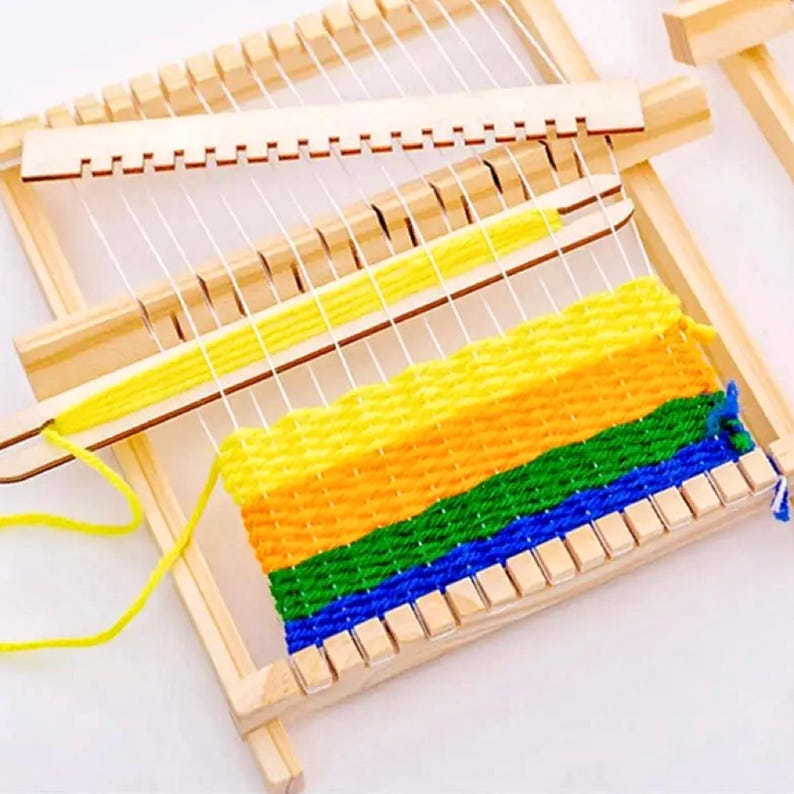May include: A wooden weaving loom with a partially completed woven piece. The woven section displays a gradient of yellow, orange, green, and blue yarn. The loom is made of light-colored wood and has white strings stretched across it.