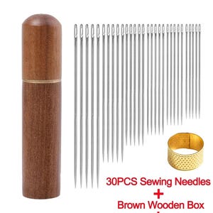 Può includere: Un kit da cucito con 30 aghi da cucito argentati, una scatola di legno marrone e un ditale dorato. Gli aghi sono lunghi e sottili, e il ditale è un anello con una superficie strutturata. Il testo dice "30PCS Sewing Needles + Brown Wooden Box + Thimble."