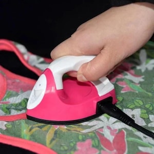 May include: A small, handheld electric iron with a bright pink body and a white handle. The iron features a black soleplate and is connected to a black power cord. It is positioned on top of a fabric garment with a green, pink, and white floral pattern.