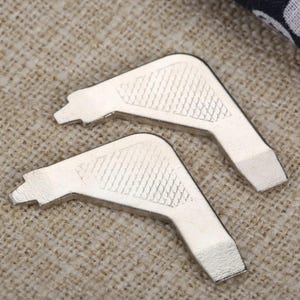 May include: Two silver-toned metal corner protectors with an L-shaped design. Each piece features a textured, cross-hatched pattern on the top surface and a smooth, flat finish on the edges, designed for securing the corners of books or boxes.
