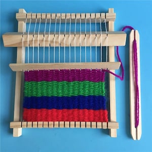 Puede incluir: Un pequeño telar de madera con hilos de urdimbre blancos y una trama parcialmente tejida de colores. La trama presenta bandas de hilo rojo, azul, verde y morado. Una lanzadera de madera con hilo morado está a la derecha.