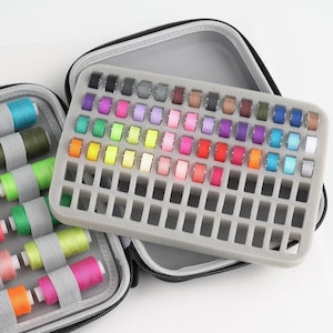 Puede incluir: Un estuche de costura gris con cremallera y un inserto de espuma lleno de coloridas bobinas de hilo. Las bobinas están dispuestas en una cuadrícula, mostrando una amplia gama de colores, incluyendo azul, verde, amarillo, rosa y morado. Bobinas adicionales son visibles en el estuche abierto.
