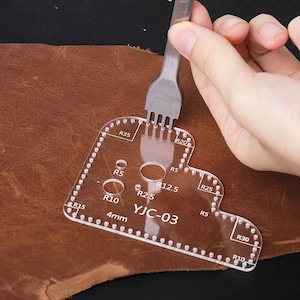May include: A clear acrylic leathercraft template with various hole sizes and rounded corners. The template has the text "YJC-03" and "4mm" printed on it. A hand holds a metal tool over the template, positioned on a piece of brown leather.